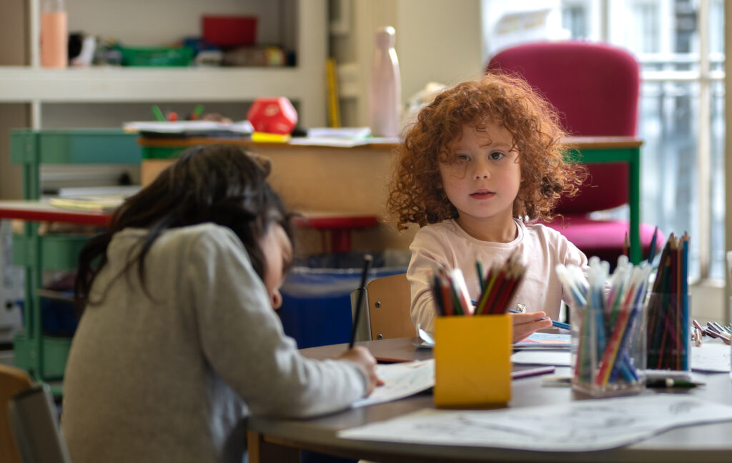 EIFA Two children are seated at a table in a bilingual classroom. One child with curly red hair looks at the camera, holding a pencil. The other child, with long dark hair, is focused on drawing. Art supplies are scattered on the table. Shelves are visible in the background.