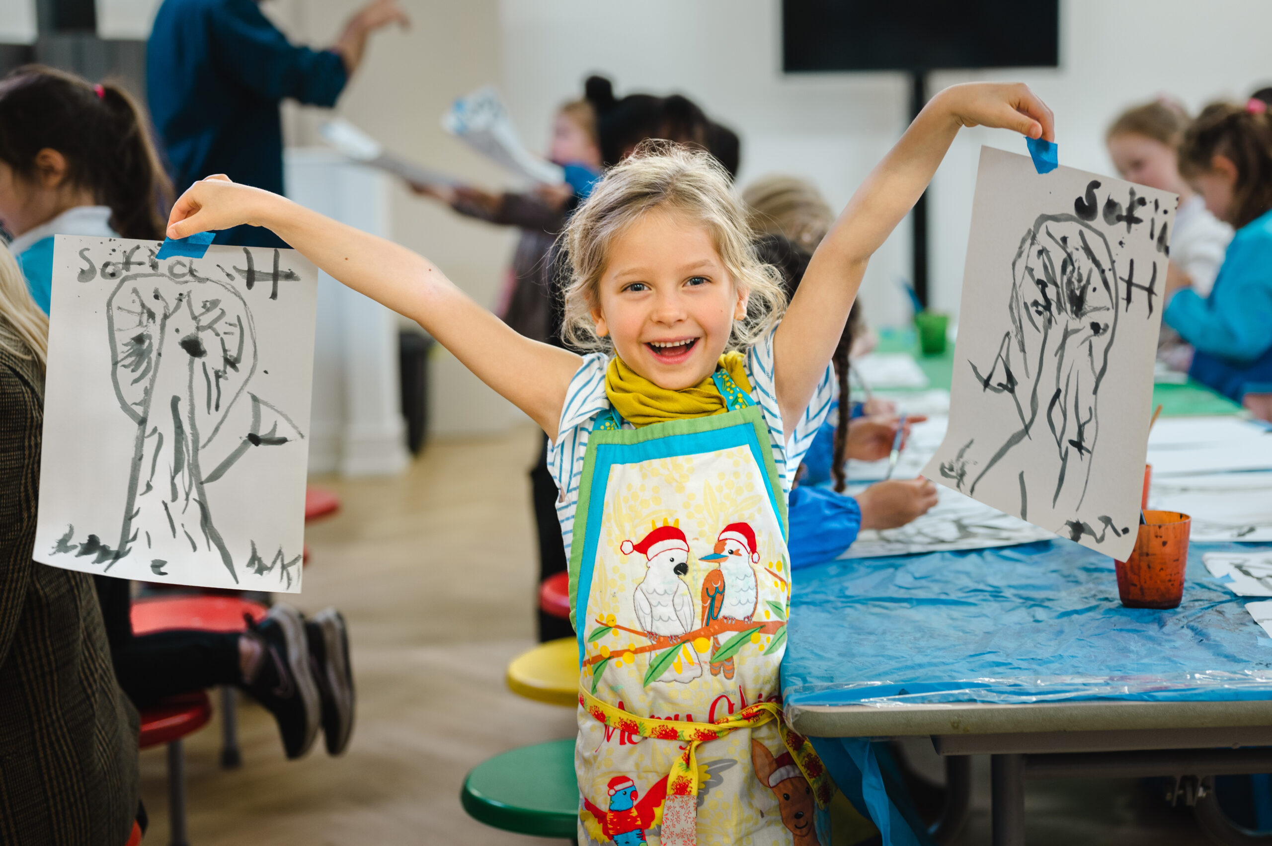 EIFA A smiling child proudly holds up two black-and-white drawings of owls at EIFA, a bilingual school. The child wears a colorful apron with a cartoon design, while in the background, other children are engaged in drawing at a table.