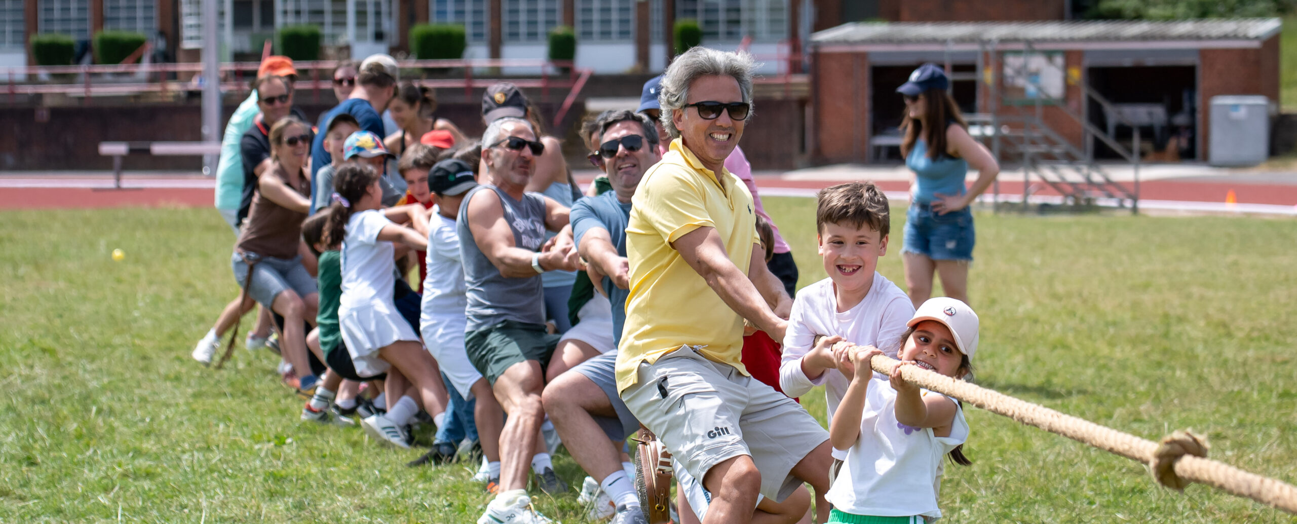 EIFA A group of people, including adults and children, are enthusiastically participating in a tug-of-war at the bilingual schools grassy field. The man in a yellow shirt and two kids upfront are smiling, while spectators watch from the background.