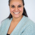 EIFA A person with long dark hair smiles at the camera, embodying a sense of bilingual charm. They are wearing a light blue textured sweater and a black top against a plain light gray background.