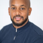 EIFA A bilingual man with a shaved head and a beard smiles softly at the camera. He is wearing a dark zip-up top against a neutral background, embodying the approachable yet professional essence of an EIFA school educator.