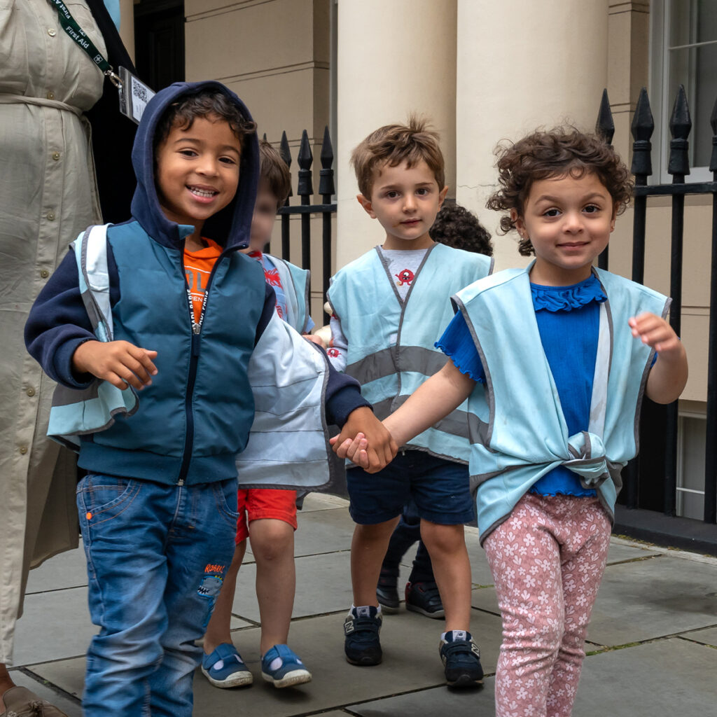 EIFA Quatre jeunes enfants marchent joyeusement ensemble sur un trottoir de Londres, vêtus de gilets de sécurité bleu clair. Ils sourient et se tiennent la main, avec une clôture en fer forgé et des colonnes en arrière-plan, se dirigeant peut-être vers leur école bilingue de lEIFA.