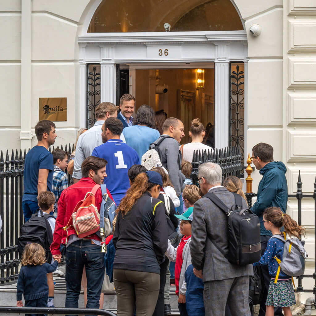 EIFA Un groupe de personnes, dont des enfants et des adultes, se rassemblent devant un bâtiment portant le numéro 36 au-dessus de la porte à Londres. Ils semblent entrer dans lEIFA, qui comporte une clôture en fer forgé noir et une plaque avec une inscription à côté de lentrée.