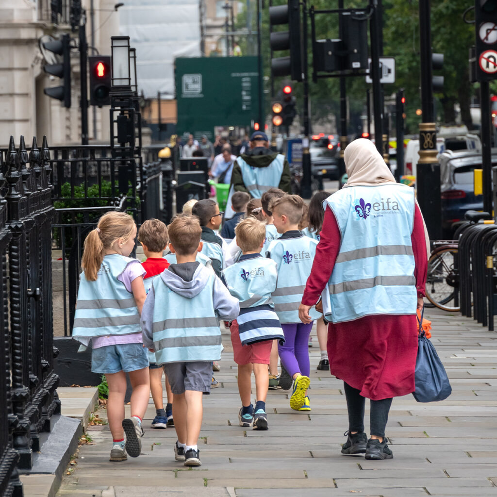 EIFA Un groupe décoliers bilingues portant des gilets réfléchissants bleu clair marchent en file indienne sur un trottoir animé de Londres, accompagnés dadultes. La scène montre leurs dos alors quils séloignent au milieu dun environnement urbain, avec des bâtiments, des vélos garés et des panneaux de signalisation.