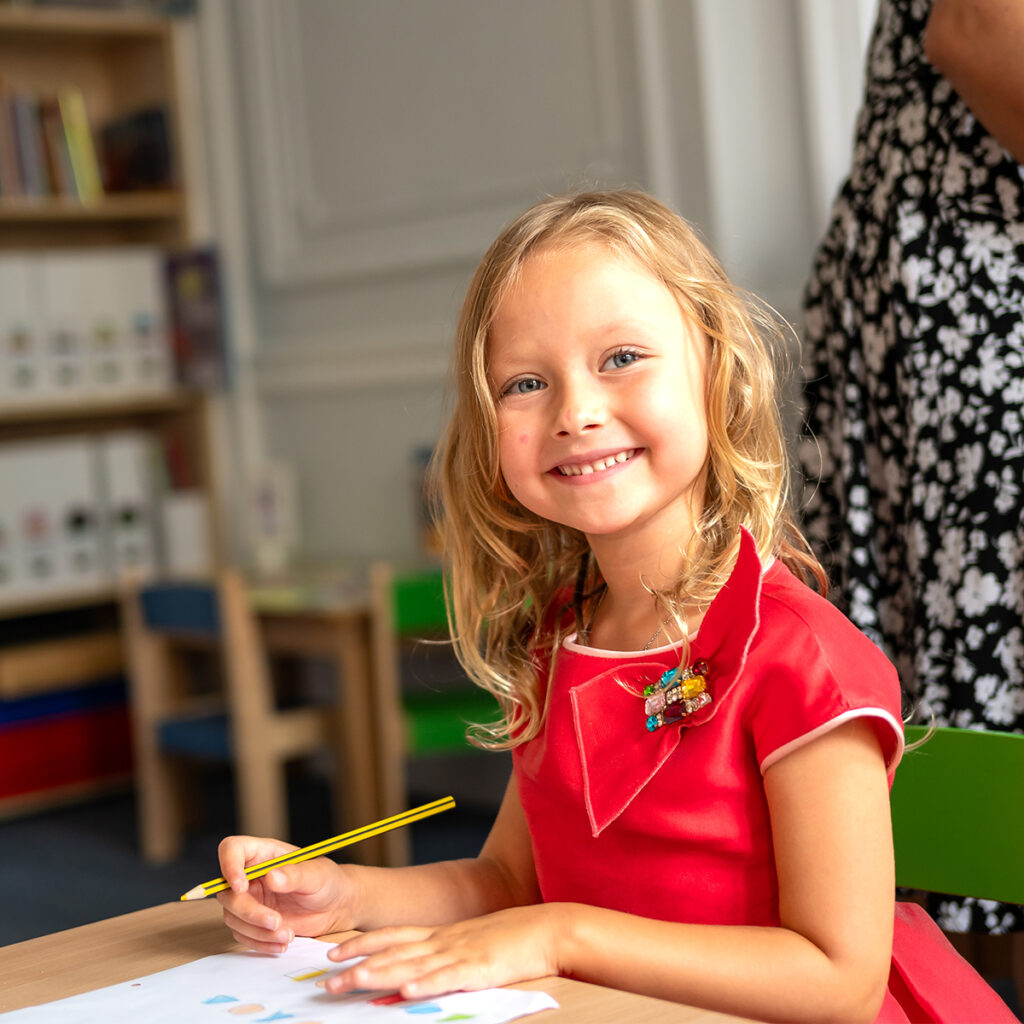 EIFA Une jeune fille aux cheveux blonds sourit tout en dessinant au crayon dans sa classe bilingue à lEIFA. Elle porte une robe rouge et est assise à une table, entourée détagères, avec une robe à motifs floraux visible en arrière-plan.