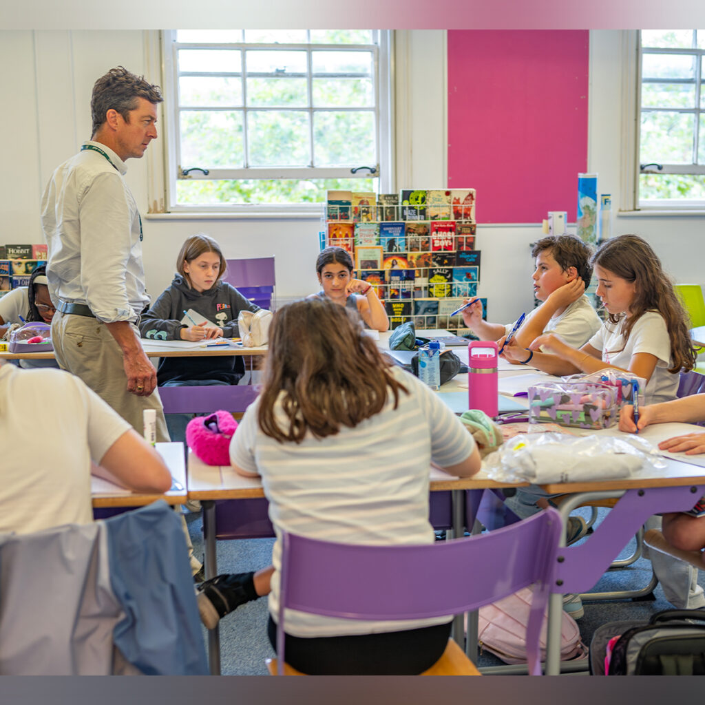 EIFA Un enseignant bilingue discute avec des élèves assis à leur bureau dans une salle de classe animée de lEIFA. Les élèves attentifs écoutent attentivement la lumière du soleil qui pénètre à travers de grandes fenêtres, illuminant les murs et les décorations colorées.