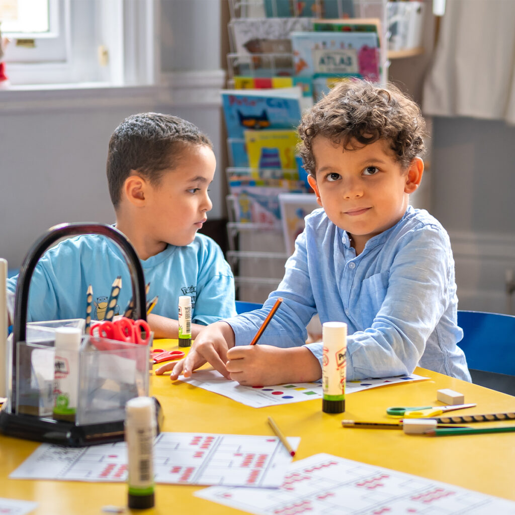 EIFA Deux jeunes garçons sont assis à une table couverte de fournitures artistiques, notamment des bâtons de colle, des marqueurs et du papier de couleur. Lun des garçons sourit à la caméra tout en tenant un crayon, et lautre regarde sur le côté. Les étagères remplies de livres reflètent les riches ressources de leur école bilingue dans le Londres dynamique.