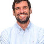 EIFA A bilingual man with a beard, wearing a light blue striped shirt, is smiling against a plain white background, embodying the diverse community of EIFA School.