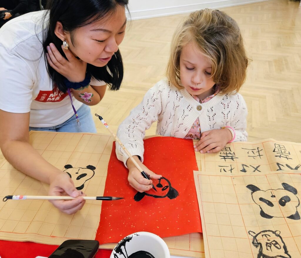 EIFA An adult helps a young girl paint a panda face on red paper using a brush. Calligraphy sheets with pandas and Chinese characters are spread on a table in front of them.