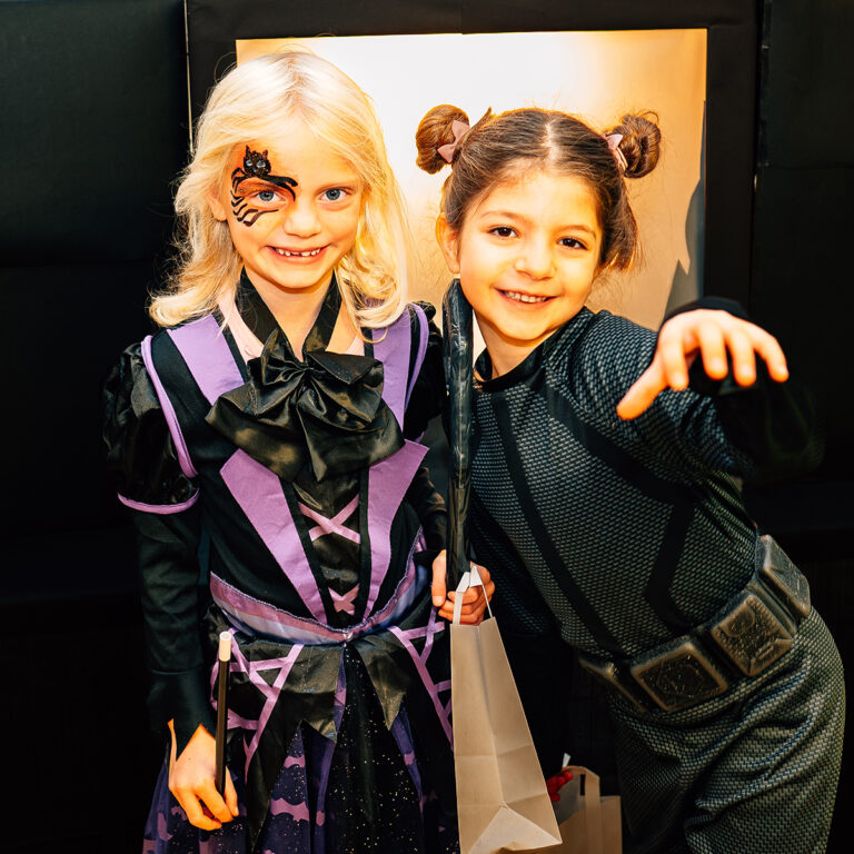 EIFA Two children from EIFA in London are smiling in Halloween costumes. The blonde child, sporting purple and black with face paint, stands next to the brunette, who dons a black outfit with playful pigtails and gestures joyfully toward the camera, showcasing their bilingual spirit.