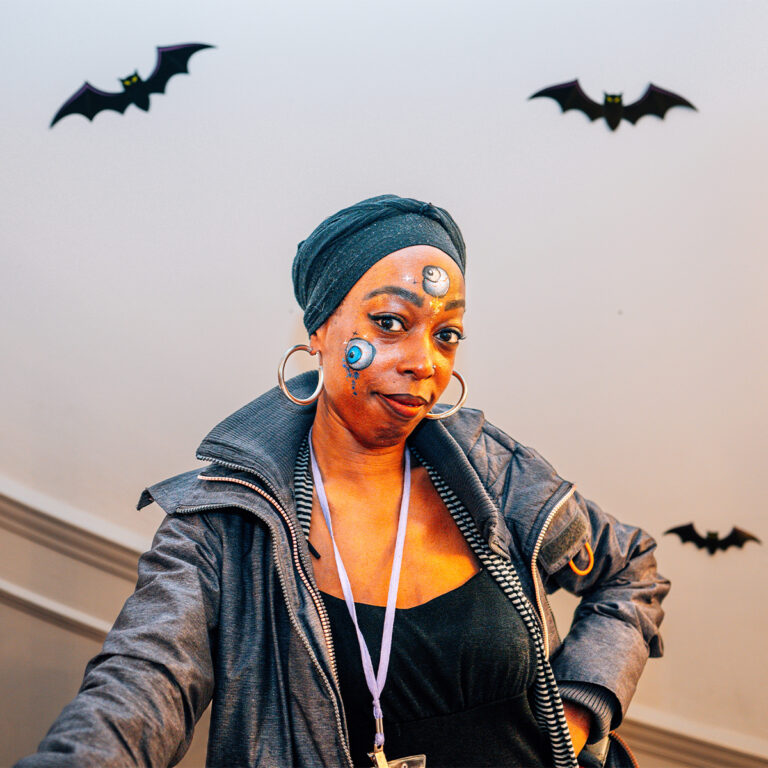 EIFA A person wearing a headscarf and hoop earrings stands indoors with a confident expression. Face paintings of planets adorn their face, complementing the jacket over their black top. Decorative black bat cutouts are displayed on the wall behind them, evoking an artistic vibe reminiscent of an EIFA school project.