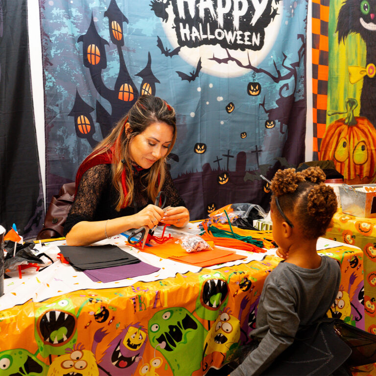 EIFA At a bilingual school, a woman assists a child with a Halloween craft project at a festive table. The backdrop features Halloween decorations with pumpkins and ghosts, while the tablecloth is adorned with colorful cartoon monsters.
