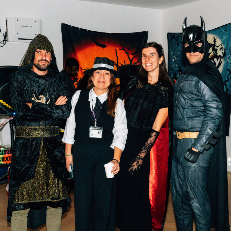 EIFA In a room adorned with Halloween decorations and banners, four people in costumes—a medieval knight, a person in a gangster outfit with a cup, a woman in a Victorian-style dress, and someone dressed as Batman—gather at the EIFA school in London, embracing the festive spirit.