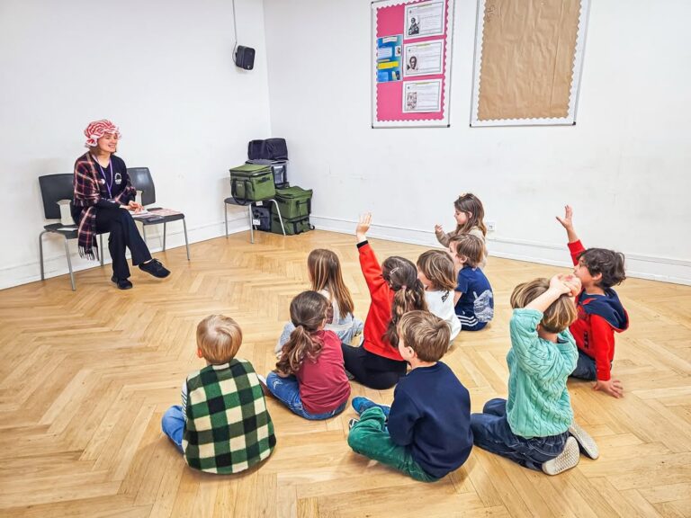EIFA A woman in a red bonnet and checked shirt sits on a chair, reading to young children seated on the floor with hands raised during International Open Day in a classroom with wooden floors and bulletin boards.