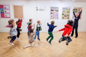 EIFA Un groupe d’enfants saute et joue joyeusement dans une salle de classe spacieuse au sol en bois, tandis qu’un adulte les guide dans une activité énergique de la Journée mondiale de l’alimentation près des tableaux d’affichage colorés sur les murs. EIFA Un groupe d'enfants saute et joue joyeusement dans une salle de classe spacieuse au sol en bois, tandis qu'un adulte les guide dans une activité énergique de la Journée mondiale de l'alimentation près des tableaux d'affichage colorés sur les murs.