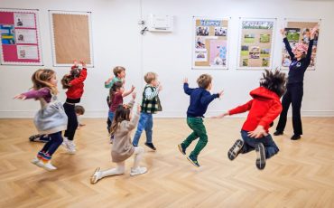 EIFA A group of children joyfully jump and play in a brightly lit classroom with wooden floors, while an adult leads them in an energetic activity near wall displays during International Open Day.