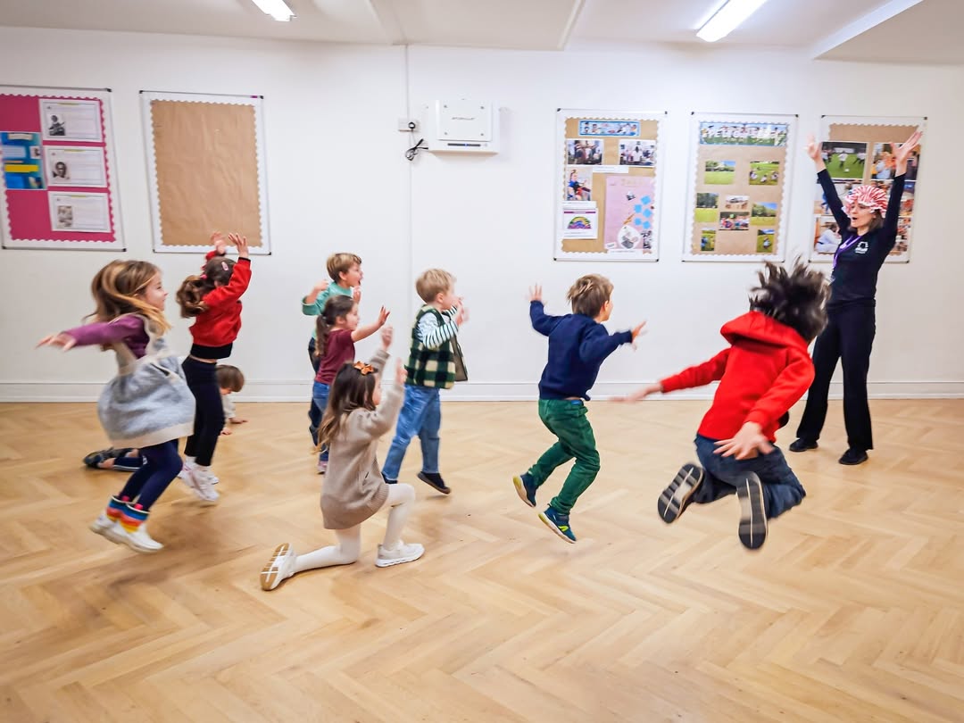 EIFA A group of children joyfully jump and play in a brightly lit classroom with wooden floors, while an adult leads them in an energetic activity near wall displays during International Open Day.