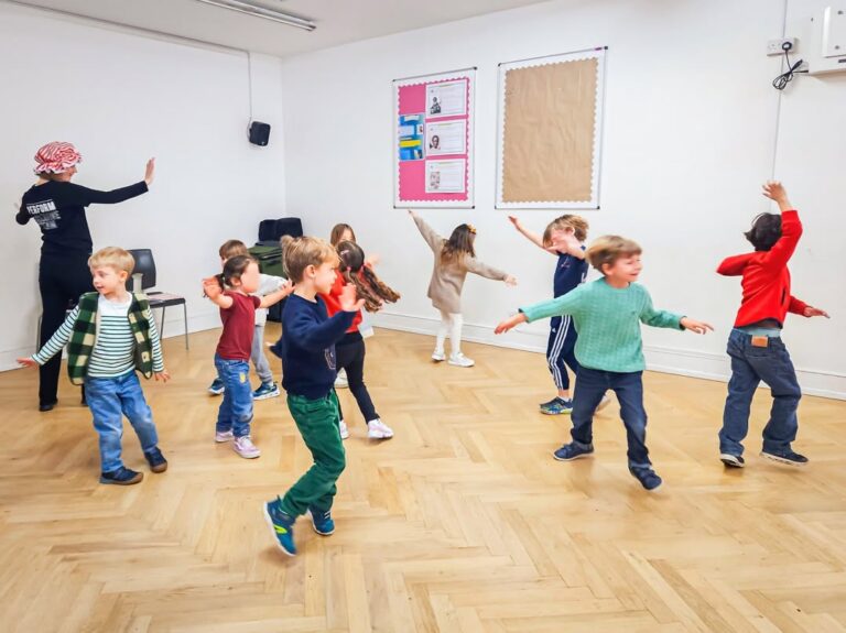 EIFA A group of young children dance and play in a brightly lit classroom with a wooden floor during International Open Day, following an adult instructor who is raising her hand. The children appear happy and engaged.