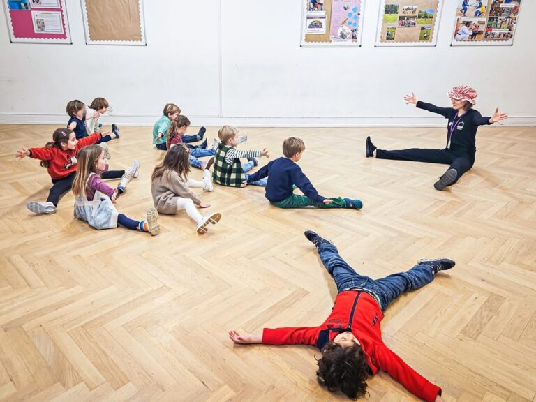 EIFA A group of children sits on a wooden floor in a circle with an adult leading them, arms stretched wide, during International Open Day. One child lies on the floor with arms and legs spread out. Colorful posters are displayed on the wall behind them.