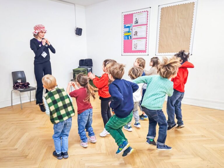 EIFA A group of young children excitedly follow a woman wearing a red hat and playing a recorder in a classroom with wooden floors and bulletin boards, celebrating International Open Day.