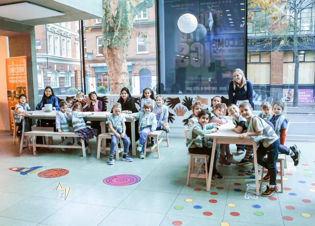 EIFA Un groupe d'enfants et quelques adultes sont assis autour de tables dans une salle de classe moderne et lumineuse dotée de grandes fenêtres. Certains enfants sourient à la caméra, tandis que d'autres parlent ou travaillent à des activités. Des formes colorées décorent le sol.