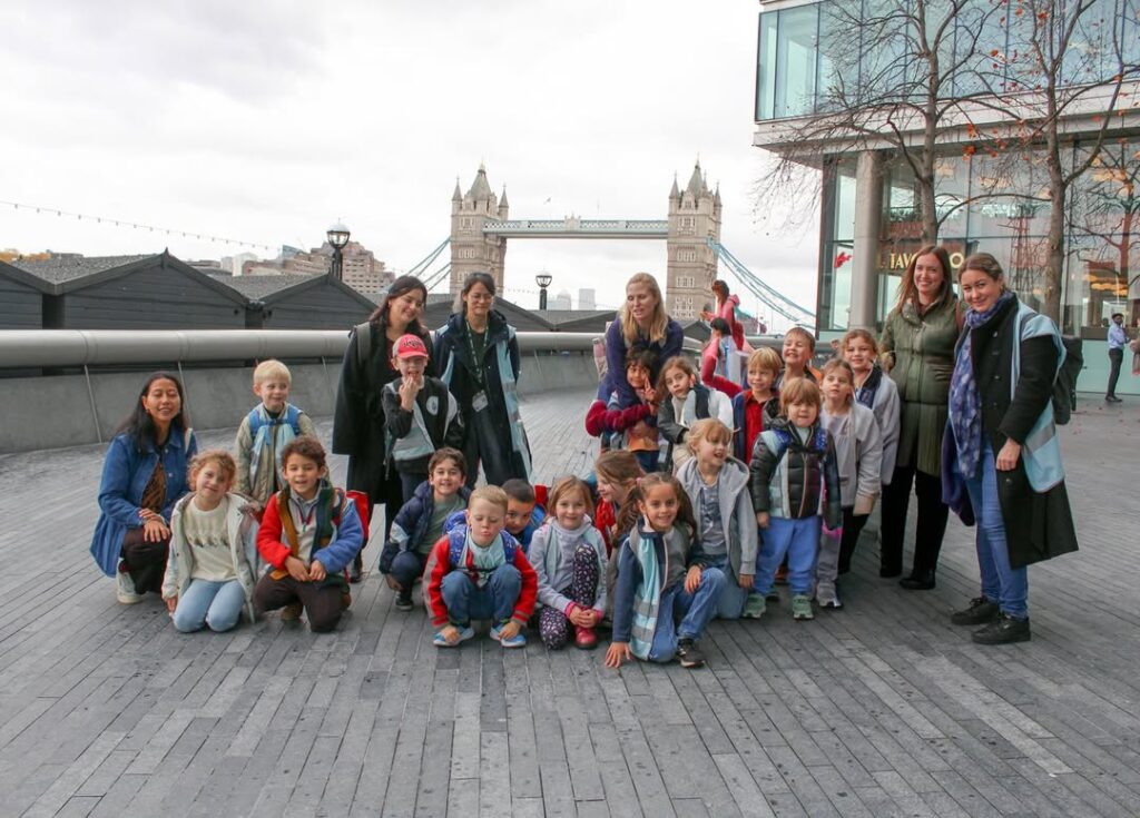 EIFA Un groupe d'enfants et d'adultes pose pour une photo en plein air près du Tower Bridge à Londres. Les enfants sont assis et debout, souriant à l'appareil photo, avec des bâtiments modernes et le pont emblématique en arrière-plan.