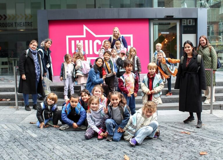 EIFA A group of children and several adults pose and smile outside a building with a large pink sign. The children appear cheerful, some kneeling in front, while the adults stand behind them.