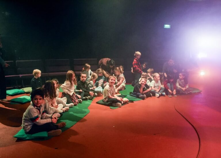 EIFA A group of young children sit on green mats on an orange floor in a dimly lit room, watching something off-camera. Some children are chatting or looking around, while a few adults are nearby in the background.