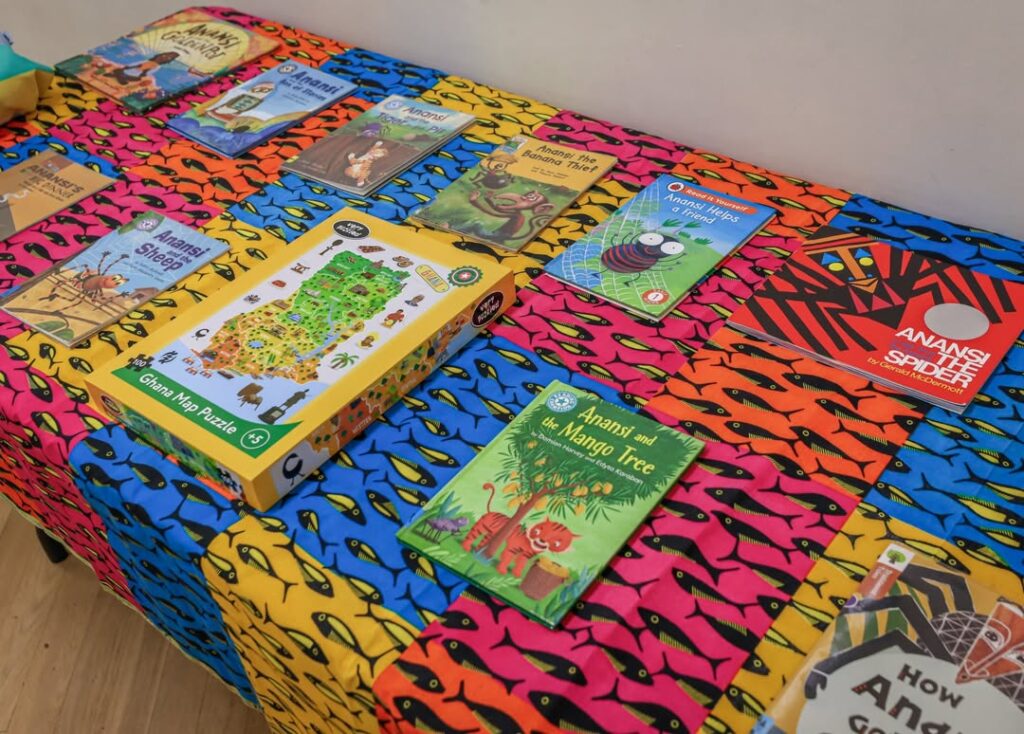 EIFA Une table colorée recouverte d'un tissu aux motifs éclatants présente divers livres pour enfants et un puzzle représentant une carte du Ghana, avec des titres comme "Anansi l'araignée" et "Anansi et le manguier".