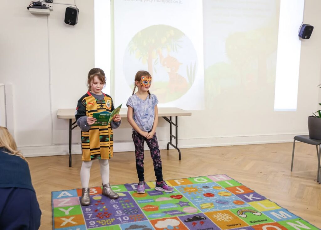 EIFA Deux jeunes enfants se tiennent devant une image d'histoire projetée dans une salle de classe. L'un d'eux tient un livre et lit, tandis que l'autre porte un masque d'animal. Un tapis d'alphabet est posé sur le sol devant eux.