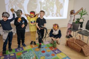 EIFA Cinq enfants portant des masques et des costumes d'animaux sont debout sur un tapis alphabétique coloré et posent pour une photo. Derrière eux se trouve une projection d'animaux de la jungle. Un autre enfant arrose une plante sur la droite. Des accessoires et des paniers sont posés sur le sol.