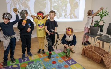EIFA Five children wearing animal masks stand on a colorful alphabet mat, posing with props in front of a screen showing animal illustrations; one child to the right is leaning toward a plant, also in costume.