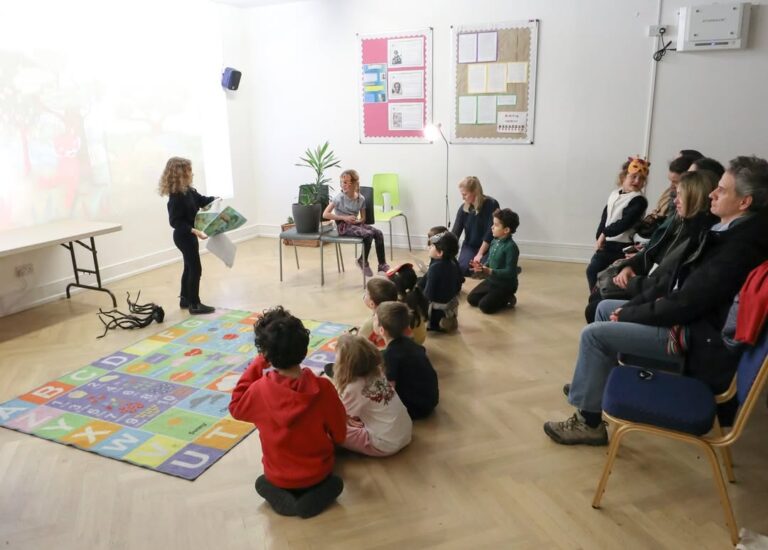 EIFA A young child stands and reads from a book to a group of children sitting on a colorful alphabet mat, while adults watch from chairs along the wall in a classroom setting.