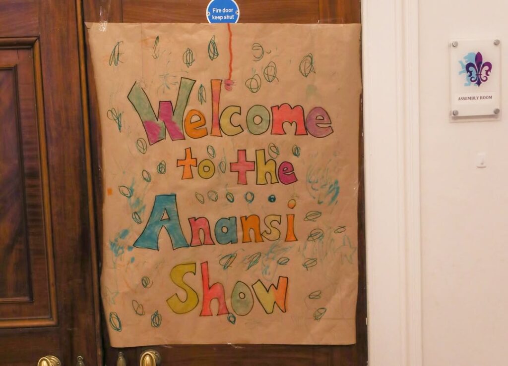 EIFA Un panneau fait à la main, sur lequel on peut lire Welcome to the Anansi Show avec des lettres colorées et des gribouillis en forme de feuilles, est collé sur une porte en bois près d'une plaque intitulée ASSEMBLY ROOM (salle d'assemblée).
