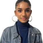 EIFA Une jeune femme aux cheveux tirés en arrière, portant de grandes boucles d'oreilles, un col roulé bleu et une veste en jean, se tient sur un fond blanc uni et regarde l'appareil photo.