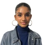 EIFA A young woman with dark hair tied back, wearing large hoop earrings, a blue turtleneck, and a denim jacket, stands against a plain white background.