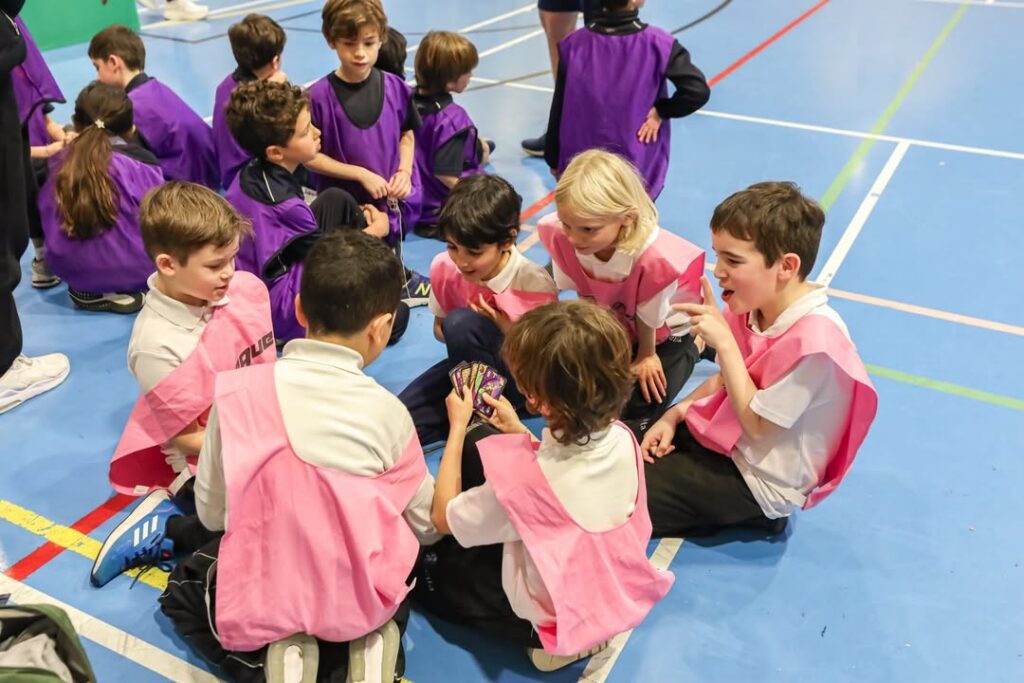 EIFA Un groupe d'enfants portant des bavoirs roses sont assis en cercle sur le sol d'un gymnase et regardent ensemble des cartes. D'autres enfants portant des bavoirs violets sont rassemblés à l'arrière-plan. Le décor est lumineux et animé.