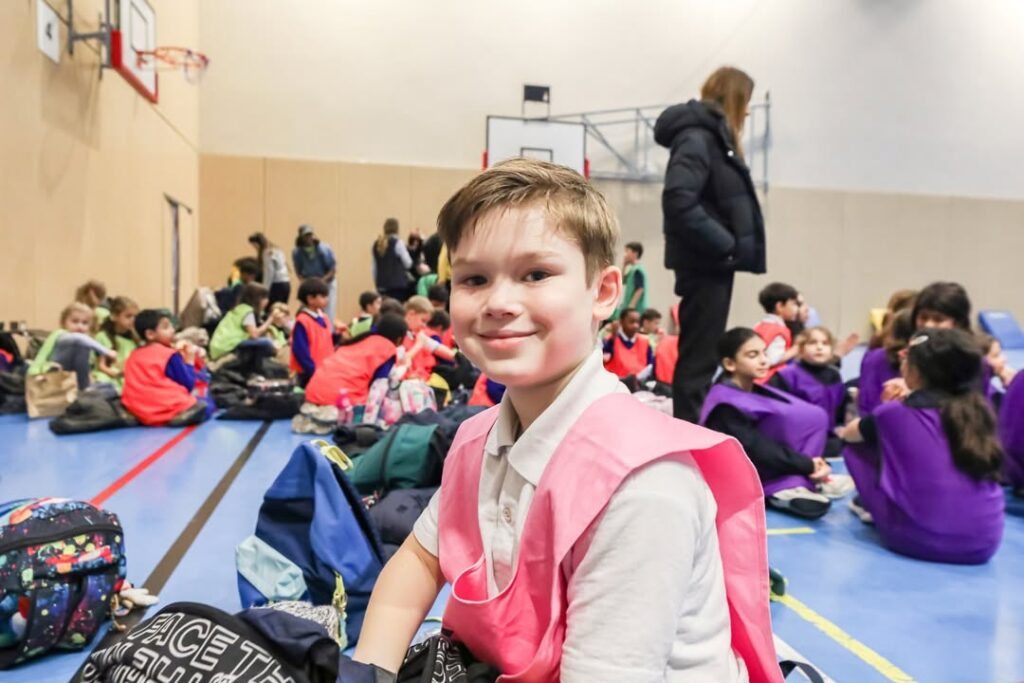 EIFA Un garçon portant un dossard de sport rose est assis sur le sol d'un gymnase et sourit à la caméra. Autour de lui, d'autres enfants portant des dossards colorés sont rassemblés en groupes, avec des sacs à dos éparpillés à proximité. Un panier de basket-ball est visible à l'arrière-plan.