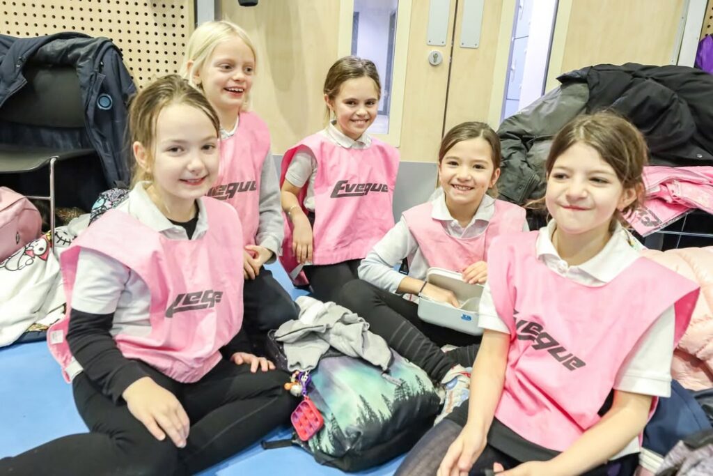 EIFA Cinq jeunes filles portant des maillots de sport roses sont assises ensemble sur le sol d'un gymnase et sourient à l'appareil photo. Elles portent des sacs et des vestes autour d'elles, ce qui suggère qu'elles sont à un événement sportif ou à un entraînement.