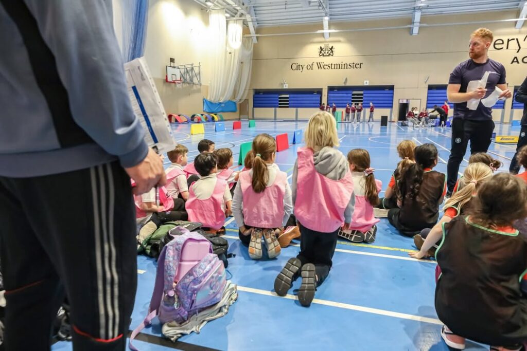 EIFA Un groupe d'enfants portant des dossards de sport roses est assis sur le sol d'un gymnase, face à un instructeur qui tient des documents. Un autre adulte se tient à proximité. Des équipements sportifs et des sacs à dos sont visibles, avec la ville de Westminster sur le mur.