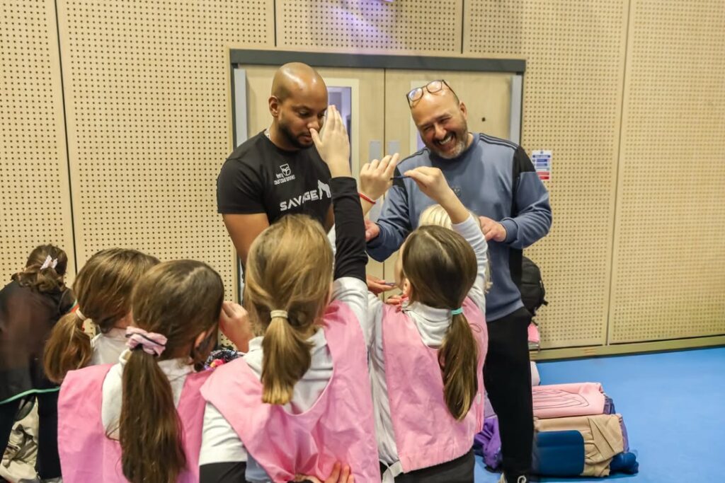 EIFA Deux hommes discutent et sourient avec un groupe de jeunes filles portant des dossards de sport roses, qui lèvent la main et font face aux hommes, dans un décor d'intérieur avec des panneaux de bois au mur.
