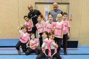 EIFA Un groupe d’enfants portant des dossards de sport roses pose avec deux entraîneurs adultes dans un gymnase. Les enfants sourient et se tiennent debout ou à genoux devant les adultes, qui sourient également. L’arrière-plan montre un mur en bois et une porte fermée. EIFA Un groupe d'enfants portant des dossards de sport roses pose avec deux entraîneurs adultes dans un gymnase. Les enfants sourient et se tiennent debout ou à genoux devant les adultes, qui sourient également. L'arrière-plan montre un mur en bois et une porte fermée.