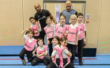 EIFA A group of young children in pink sports bibs pose and smile with two adult coaches in a gymnasium. The children are lined up in front, and the adults stand behind them, all looking happy and energetic.