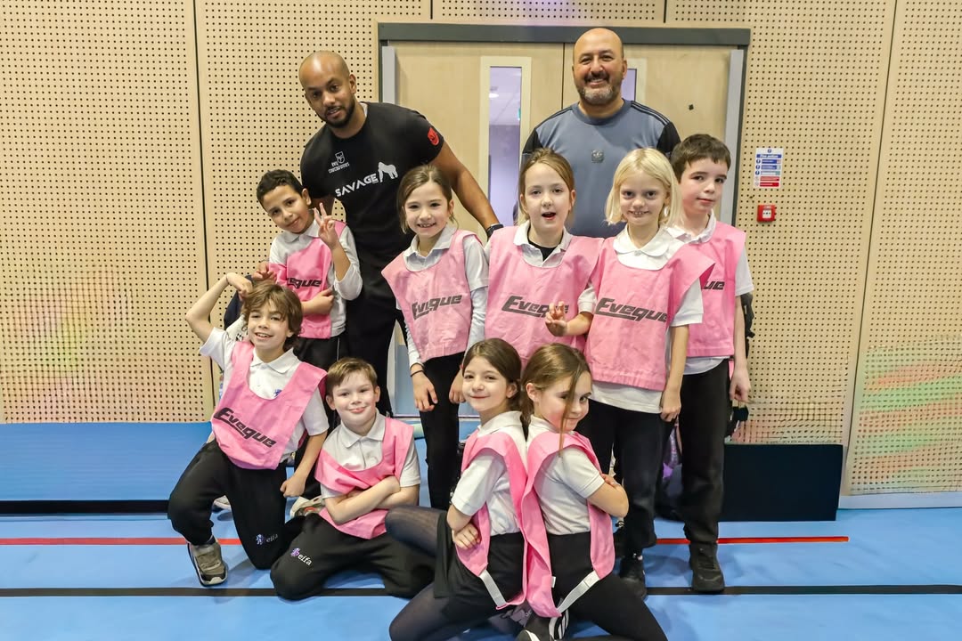 EIFA A group of young children in pink sports bibs pose and smile with two adult coaches in a gymnasium. The children are lined up in front, and the adults stand behind them, all looking happy and energetic.