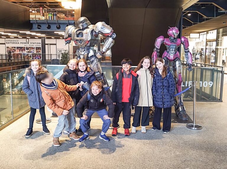EIFA A group of eight children stand smiling in front of two large robot statues, one silver and one pink, inside a modern indoor space with glass railings and various signs during International Open Day.