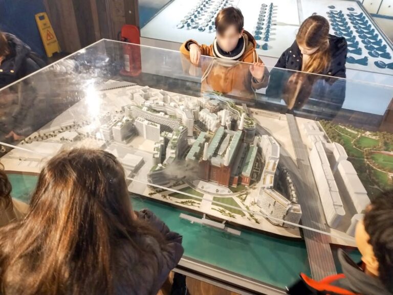 EIFA During International Open Day, a group of children observe a detailed architectural model of a city with buildings, streets, and green areas under a clear display case inside a museum or exhibition space.