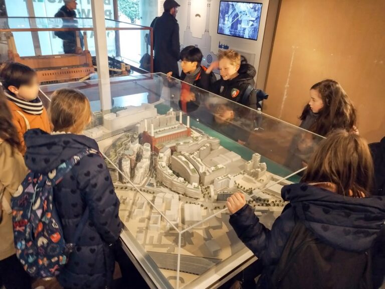 EIFA A group of children stand around a glass-covered architectural model, observing and discussing the miniature cityscape during an International Open Day at the museum or exhibition.