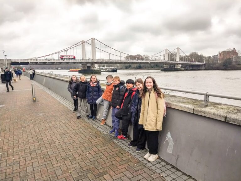 EIFA Eight children in winter clothing stand in a row, smiling near a riverside walkway during International Open Day, with a bridge, red double-decker bus, and cloudy sky in the background as others stroll along the path.