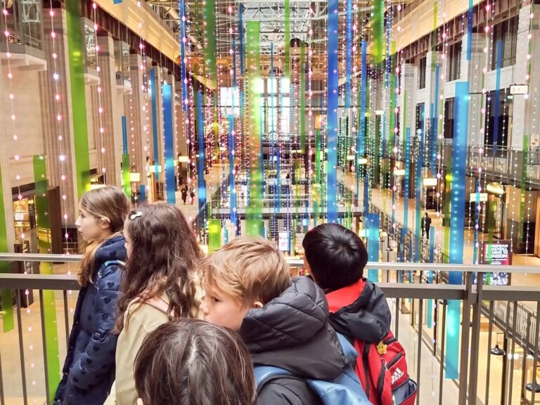 EIFA A group of children stand on an indoor balcony during International Open Day, gazing at a colorful, modern light installation made of vertical, glowing strings inside a large, bright atrium with tall windows and shops.