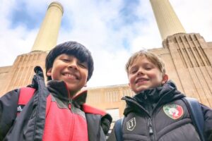 EIFA Deux garçons souriants vêtus de vestes d’hiver se tiennent à l’extérieur, avec derrière eux les hautes cheminées en briques d’un grand bâtiment. Ils regardent l’appareil photo dans un ciel bleu, capturant un moment de joie à l’occasion de la Journée mondiale de l’alimentation. EIFA Deux garçons souriants vêtus de vestes d'hiver se tiennent à l'extérieur, avec derrière eux les hautes cheminées en briques d'un grand bâtiment. Ils regardent l'appareil photo dans un ciel bleu, capturant un moment de joie à l'occasion de la Journée mondiale de l'alimentation.